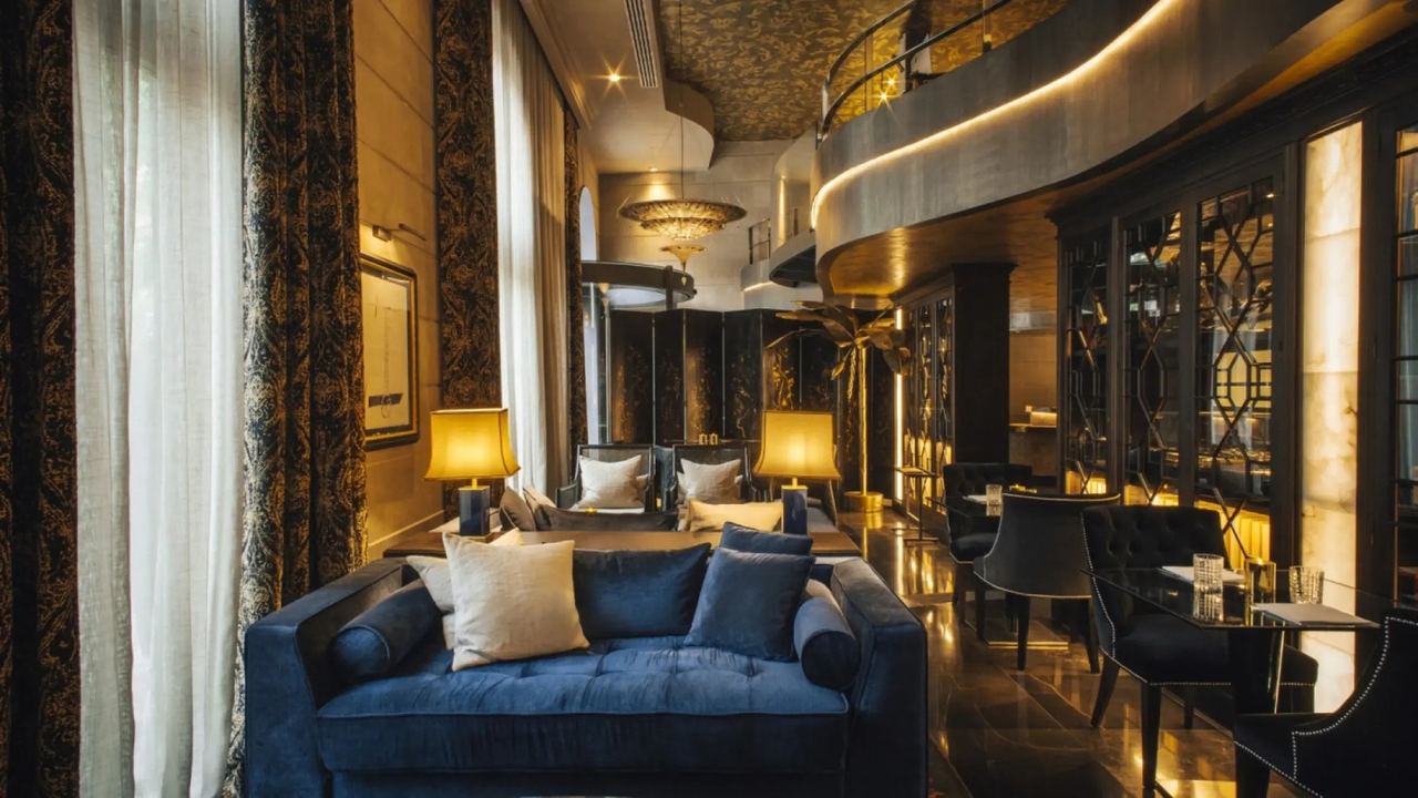 The lounge at Hotel Bagués, blue velvet sofas and lamplight beneath a gilded mezzanine