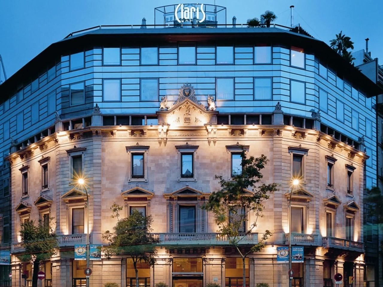 The Hotel Claris at dusk, its lit stone Palau Vedruna façade beneath a glass upper storey