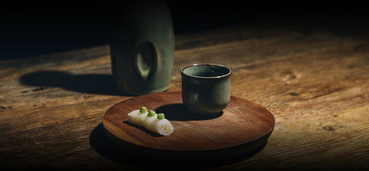 A single white bite on a wooden tray beside a sake cup at Kamikaze