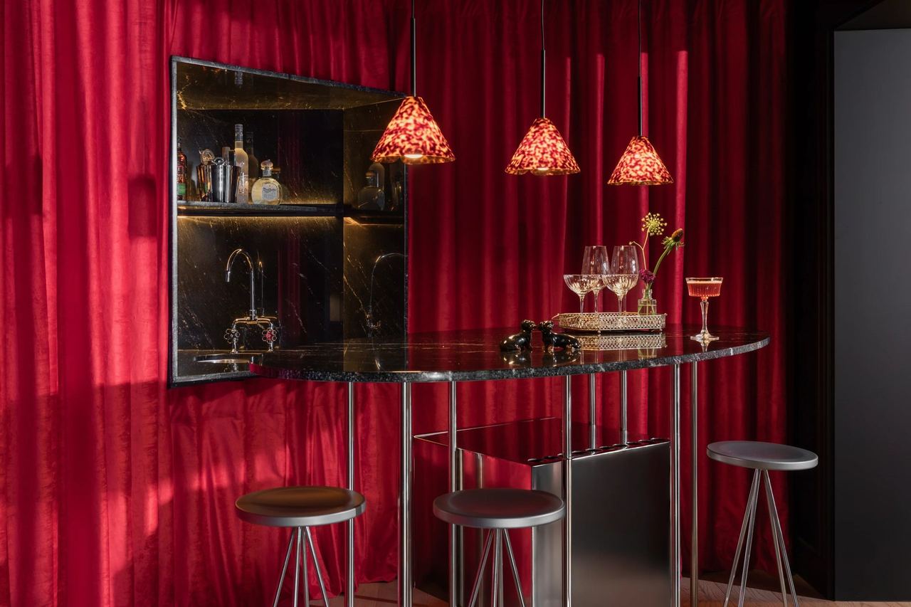 A marble bar corner with stools under red velvet curtains and glass pendants at The Wittmore